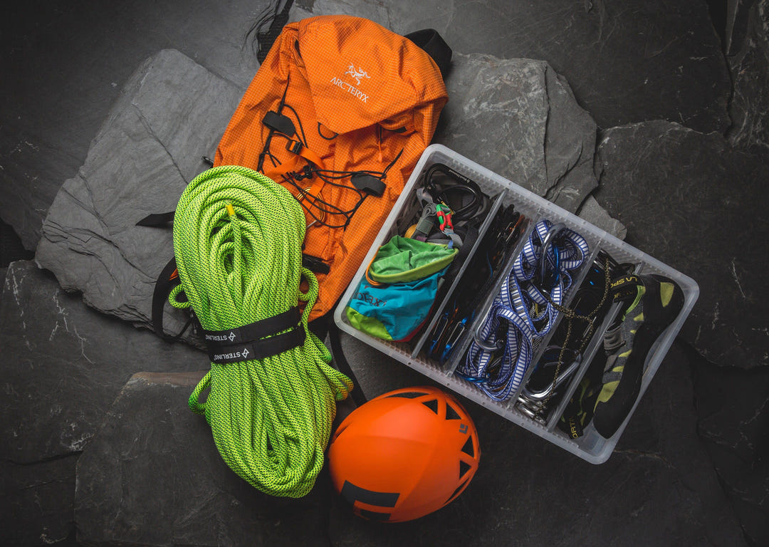 SidioCrate clear storage bin with climbing gear, lime green rope, and orange helmet on dark rocks.